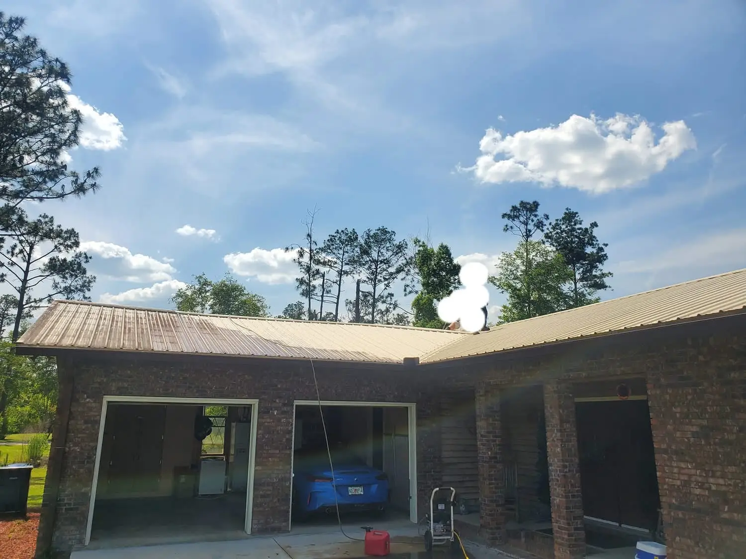 A Faith Lawn Care crew member on the metal roof of a brick Panhandle home, pressure washing it with a hose run up from the machine on the driveway